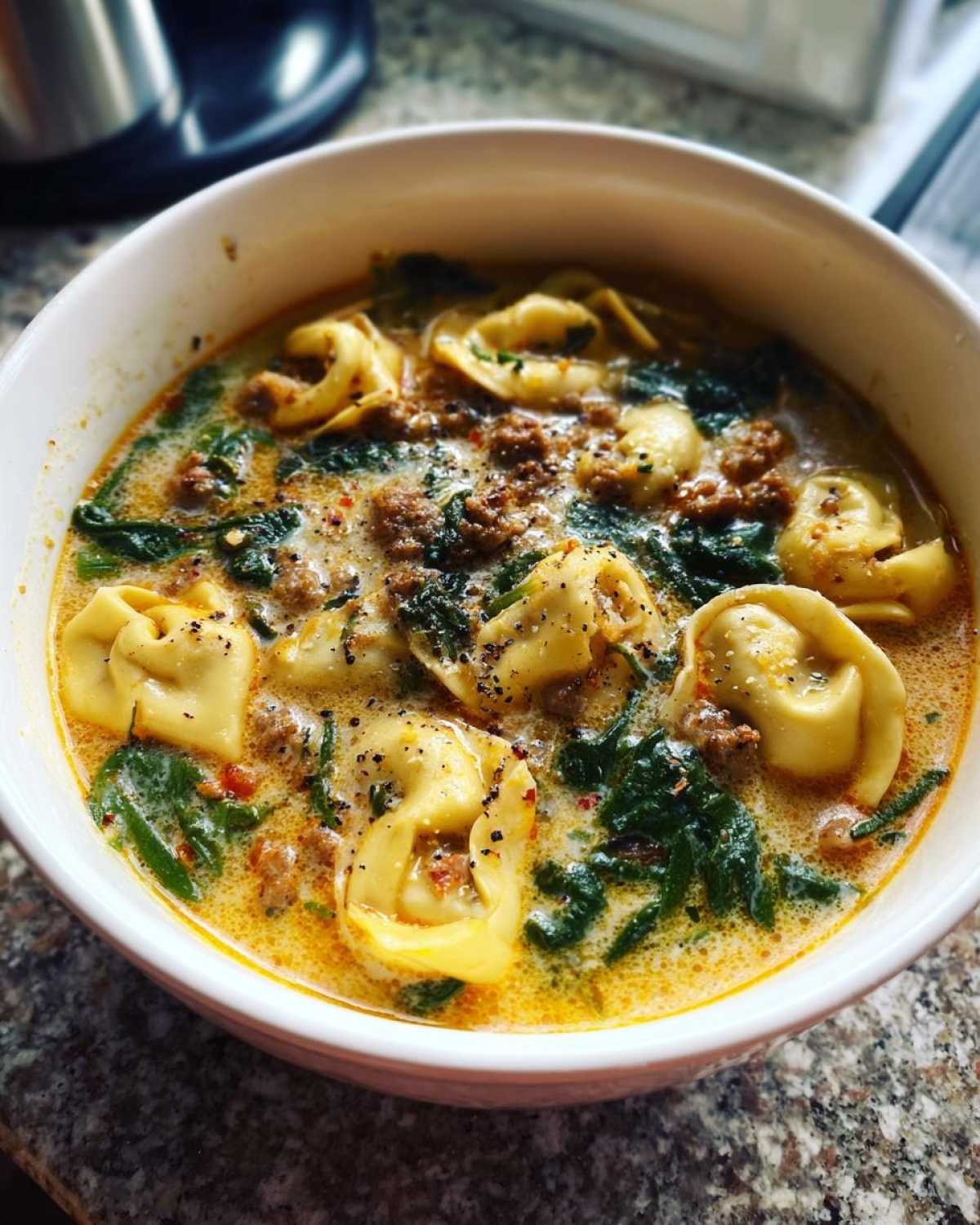 Close-up of a white bowl filled with creamy sausage tortellini soup, featuring tortellini, ground sausage, and wilted spinach.