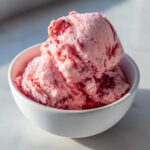 Three scoops of vibrant pink, homemade strawberry ice cream with visible fruit swirls in a small white bowl.