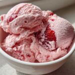 Close-up of three scoops of pink, creamy strawberry ice cream with visible chunks of real strawberries in a white bowl.