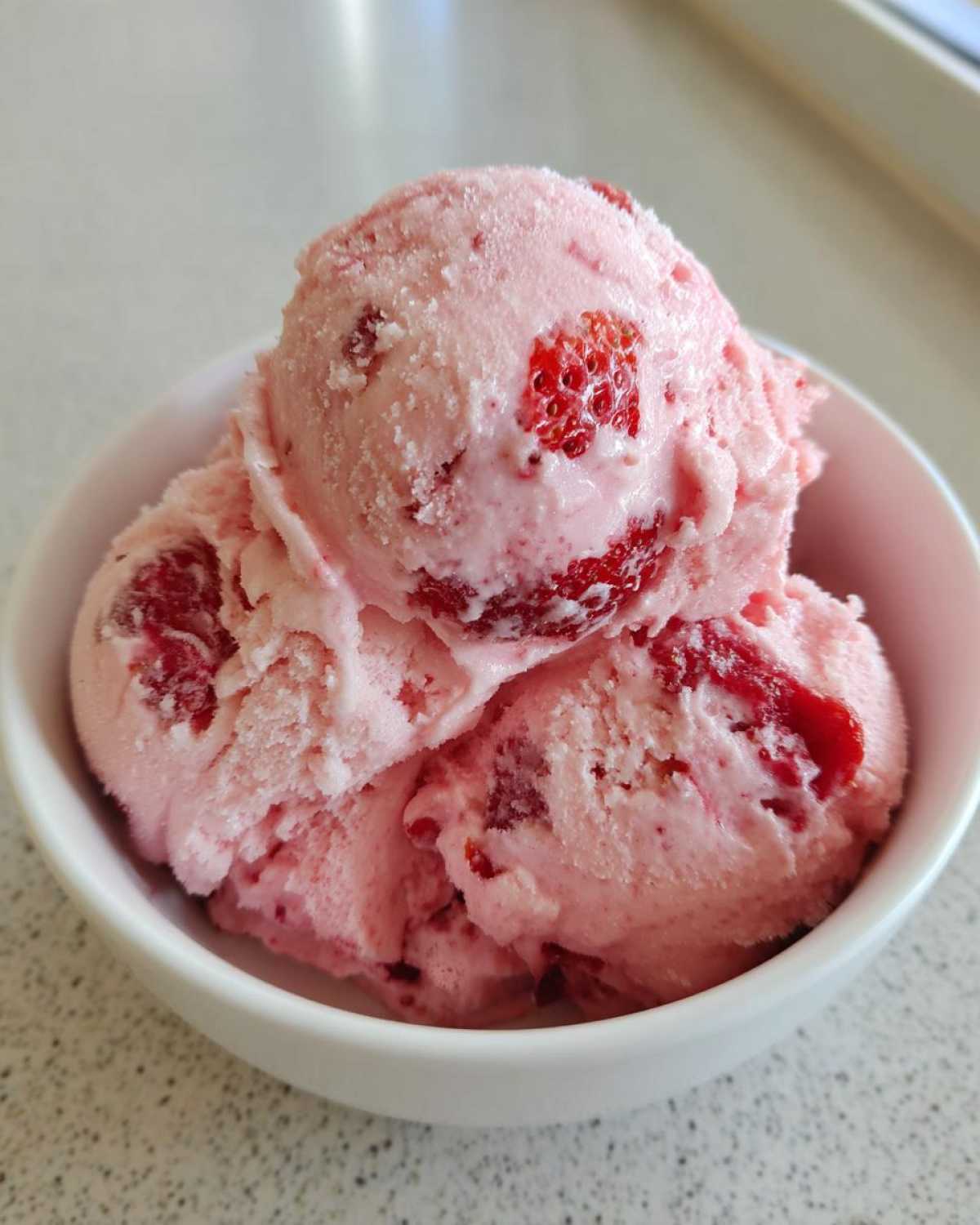 Three scoops of homemade strawberry ice cream, rich with visible strawberry pieces, served in a white bowl.