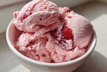 Close-up of three scoops of pink, creamy strawberry ice cream with visible chunks of real strawberries in a white bowl.