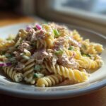 A close-up of creamy tuna pasta salad made with fusilli pasta, tuna, red onion, and green onions on a white plate.