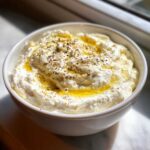 A close-up of creamy whipped feta dip in a white bowl, drizzled with olive oil and sprinkled with black pepper.
