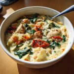 A close-up of a white bowl filled with creamy white chicken lasagna soup, featuring chicken pieces, spinach, and sun-dried tomatoes.