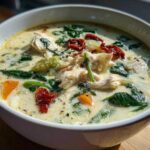 A close-up of a white bowl filled with creamy white chicken lasagna soup, featuring shredded chicken, spinach, and sun-dried tomatoes.
