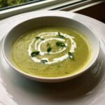 A bowl of vibrant green zucchini soup, garnished with a white cream swirl and fresh green herbs.