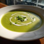 A vibrant bowl of creamy green zucchini soup, garnished with a white cream swirl and fresh microgreens.