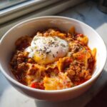 A close-up of a white bowl filled with rich crockpot lasagna soup, featuring meat sauce, pasta pieces, and topped with a poached egg and herbs.