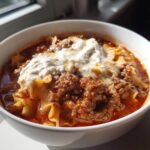 A close-up of a white bowl filled with rich crockpot lasagna soup, topped generously with ricotta cheese.