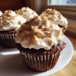 A close-up of a rich, dark chocolate hot chocolate cupcake topped with fluffy white frosting dusted with cinnamon.