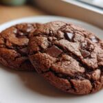 Close-up of two rich, dark brown vegan chocolate chip cookies with large chunks of melted chocolate on a white plate.