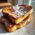 A stack of three slices of golden brown easy french toast dusted generously with powdered sugar on a white plate.