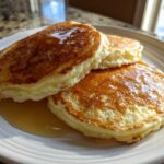 Three thick, fluffy buttermilk pancakes drizzled with golden syrup on a white plate.