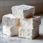 A stack of five fluffy, square homemade marshmallows heavily dusted with powdered sugar on a marble surface.