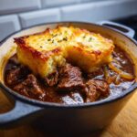 Close-up of tender beef chunks and onions in rich gravy topped with cheesy, toasted bread in a small cast iron bowl, featuring french onion beef stew.