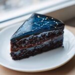 A slice of dark chocolate layer cake, a Capricorn cake, topped with a glossy blue mirror glaze featuring a gold constellation design.