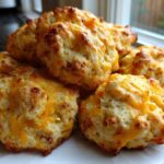 A close-up of freshly baked bisquick sausage balls piled on a white plate, topped with melted cheddar cheese.