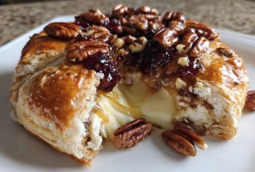 Close-up of baked brie wrapped in puff pastry, oozing cheese, topped with pecans and a sticky glaze.