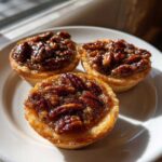 Close-up of three glistening, gooey mini pecan pies with pecan halves on top, sitting on a white plate in sunlight.