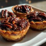 Four glistening, freshly baked mini pecan pies with caramelized pecan topping resting on a bright white serving dish.