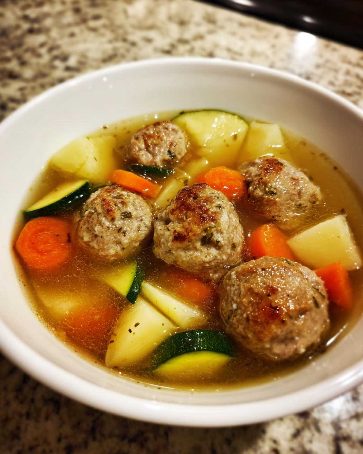A close-up of a white bowl filled with flavorful albondigas soup, featuring meatballs, carrots, zucchini, and potatoes.