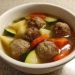 Close-up of a white bowl filled with savory albondigas soup featuring meatballs, carrots, zucchini, and potatoes.