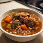 Close-up of a steaming white bowl filled with rich beef barley soup, featuring chunks of beef, carrots, and barley.