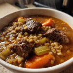 Close-up of a steaming white bowl filled with hearty beef barley soup, showing chunks of beef, carrots, celery, and barley.