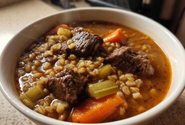 Close-up of a steaming white bowl filled with hearty beef barley soup, showing chunks of beef, carrots, celery, and barley.
