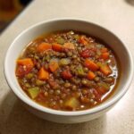 A close-up of a white bowl filled with hearty lentil soup, featuring visible lentils, diced carrots, celery, and tomatoes, seasoned with pepper.