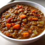 Close-up of a thick, hearty lentil soup filled with diced carrots and celery in a white bowl.