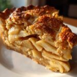 Close-up of a thick slice of homemade apple pie showing layered, spiced apples and a golden, sugared crust.