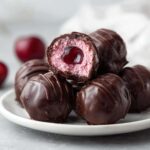 A stack of rich, dark chocolate covered cherries, one cut open showing the pink filling and whole cherry center.