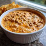 Close-up of a bubbling, creamy sausage cheese dip topped with browned sausage bits, served hot in a white bowl next to tortilla chips.