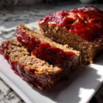 Two thick slices of juicy classic meatloaf topped with a shiny red ketchup glaze, served on a white plate.