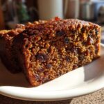 Close-up of a moist, dark slice of fruit cake loaded with dried fruit, sitting on a white plate.