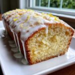 Close-up of a freshly baked lemon loaf cake with thick white icing dripping down the sides and topped with bright yellow lemon zest.