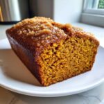 A close-up view of a freshly baked loaf of moist pumpkin bread with a sugary, glazed crumb topping on a white plate.