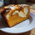 A thick slice of moist pumpkin cream cheese bread, showing the orange pumpkin cake swirled with a white cream cheese center and topped with glaze.