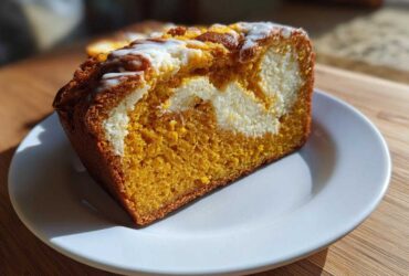 A thick slice of moist pumpkin cream cheese bread, showing the orange pumpkin cake swirled with a white cream cheese center and topped with glaze.