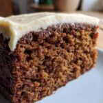 Close-up of a moist slice of dark brown gingerbread cake topped with thick cream cheese frosting.