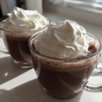 Two clear glass mugs filled with rich, dark whipped hot chocolate, generously topped with fluffy white whipped cream.