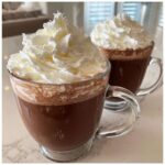 Two clear glass mugs filled with rich, dark whipped hot chocolate topped generously with fluffy white whipped cream.
