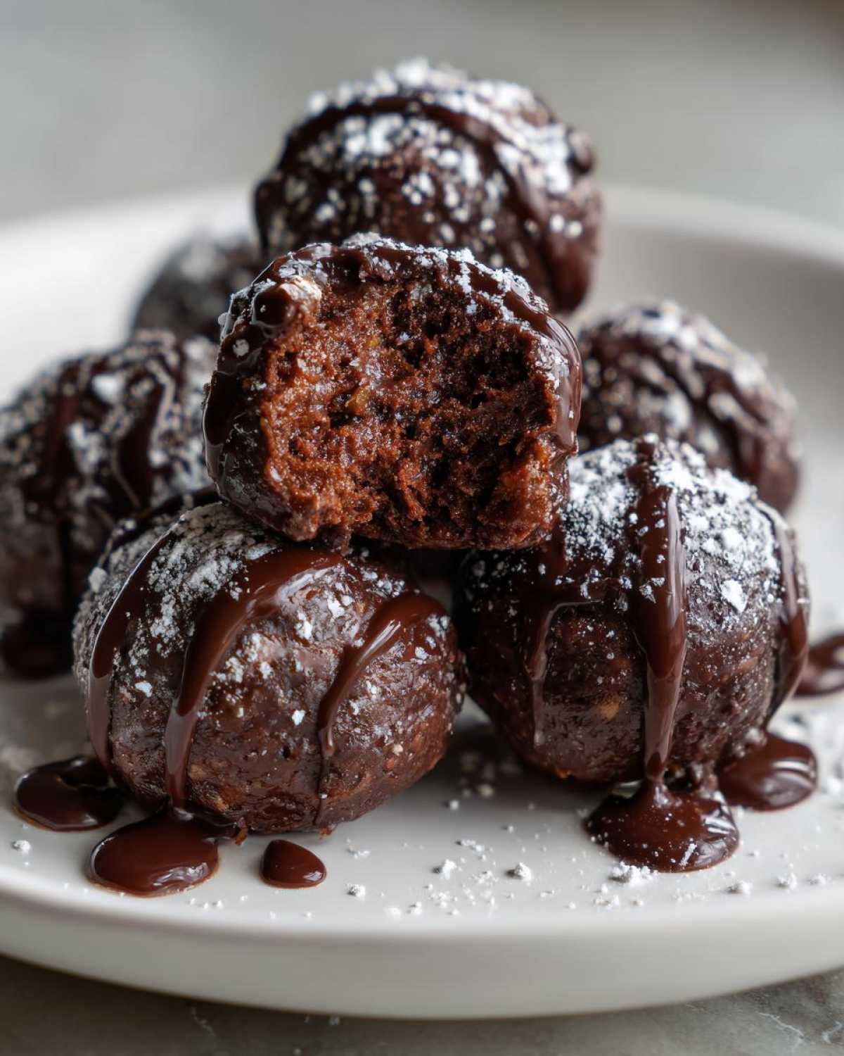 Close-up of several rich, dark chocolate pecan pie balls stacked on a plate, drizzled with chocolate sauce and dusted with powdered sugar.