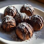 Six rich, round pecan pie balls drizzled with dark chocolate and dusted with powdered sugar on a white plate.
