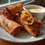 A plate of crispy, golden brown peach cobbler egg rolls, one cut open showing the fruit filling, next to a ramekin of cream sauce.