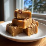 Stack of rich, brown penuche fudge squares topped with chopped pecans on a white plate.