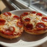 Two freshly baked pepperoni pizza bagels topped with melted cheese and herbs sitting on a white plate.