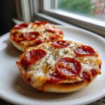 Two freshly baked pepperoni pizza bagels topped with melted mozzarella and dried herbs served on a white plate.