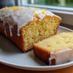 A golden-brown lemon loaf cake, partially sliced, topped with white glaze and lemon zest.